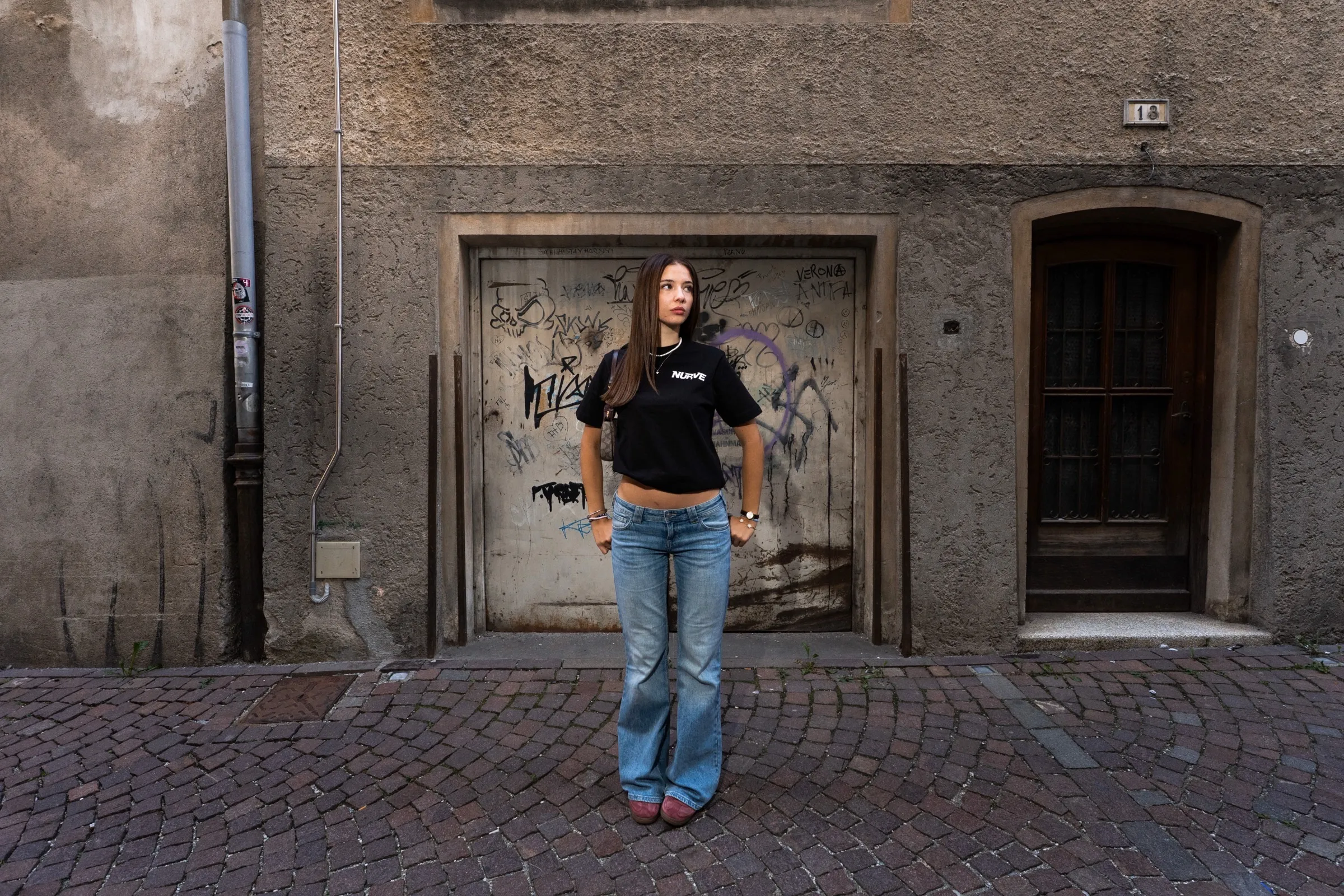 Model in schwarzem NURVE-T-Shirt und blauer Jeans vor einer grauen Hausfassade mit Türen und Graffiti